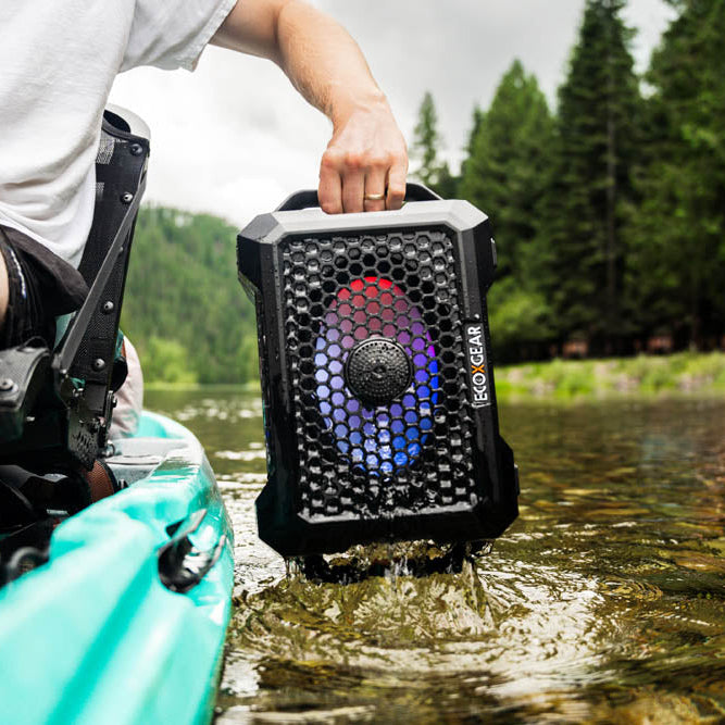 Person holding an ECOXGEAR Defender waterproof speaker in a kayak by a forested area